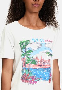 Hvid bomulds t-shirt med et farverigt grafisk motiv af palmer, blomster og en havudsigt med en kystby i baggrunden.