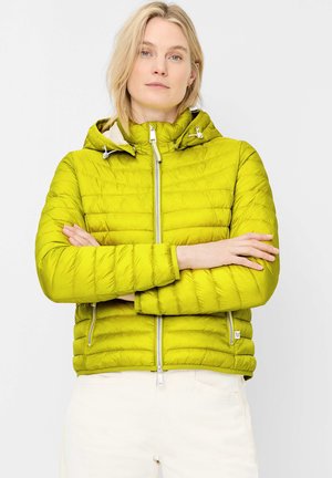 Woman with short blonde hair wearing lime green quilted jacket with hood, arms crossed, standing against white background.