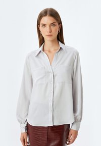 Helles graues Langarmshirt mit durchgehender Knopfleiste, zwei Brusttaschen und einem lockeren Schnitt. Glattes Material mit einem leicht schimmernden Effekt.