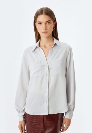 Helles graues Langarmshirt mit durchgehender Knopfleiste, zwei Brusttaschen und einem lockeren Schnitt. Glattes Material mit einem leicht schimmernden Effekt.