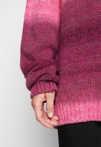 Main reposant le long du corps, portant un pull en maille dégradé du rose au magenta foncé avec des poignets et un ourlet côtelés, sur un pantalon noir.