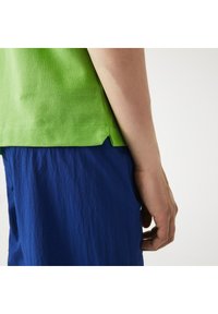 Grön texturerad kortärmad skjorta med en något längre baksida, matchad med blå skrynkliga shorts. Närbild av sidan och handen.