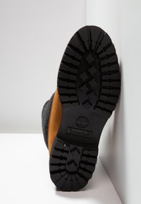 Semelle de botte Timberland avec un motif de bande de roulement en caoutchouc noir, des accents en daim marron et une tige en tissu gris texturé.