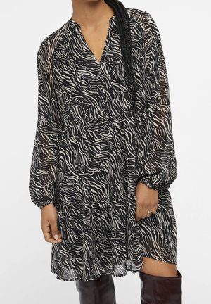 Femme portant une robe à manches longues imprimée zèbre noir et blanc avec des poignets froncés et des bottes hautes marron foncé.