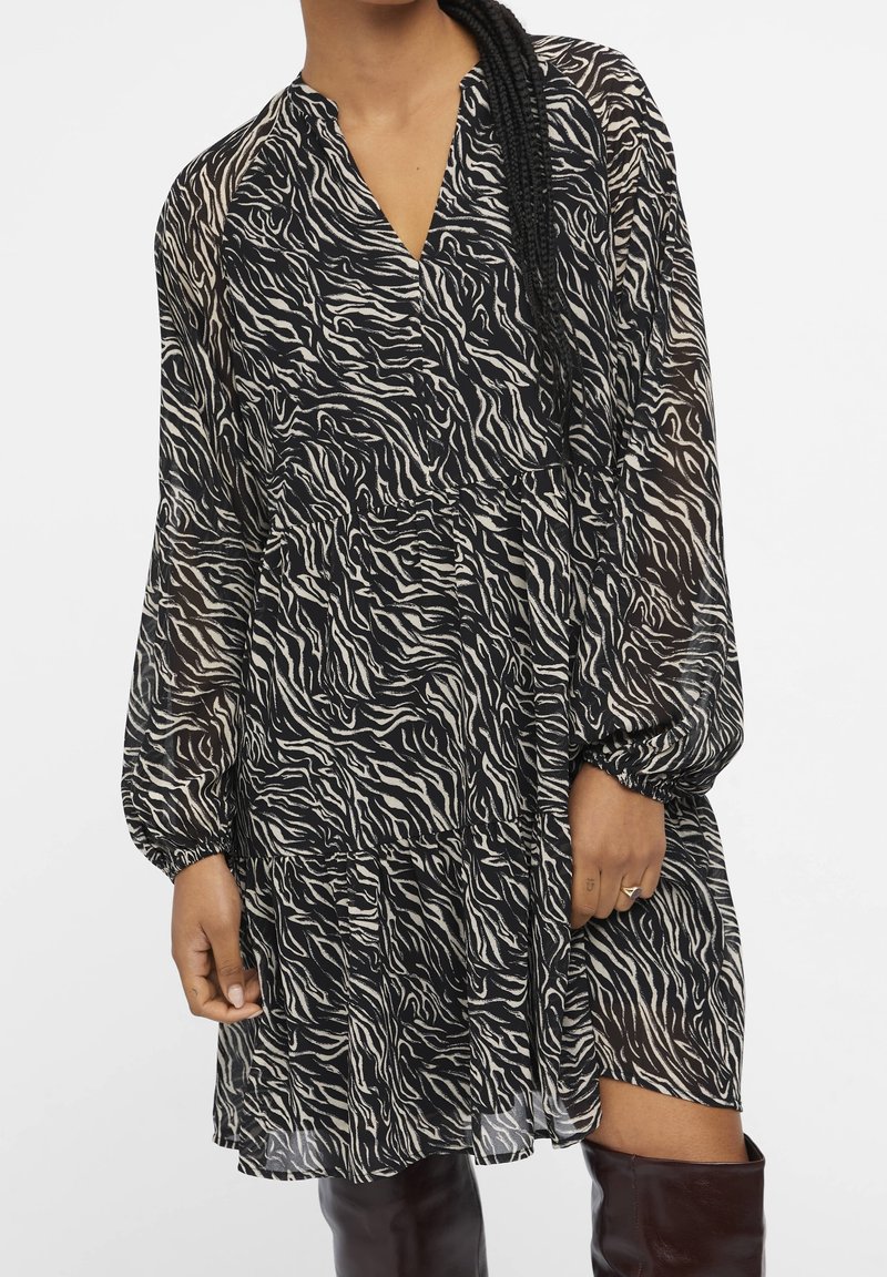 Femme portant une robe à manches longues imprimée zèbre noir et blanc avec des poignets froncés et des bottes hautes marron foncé.