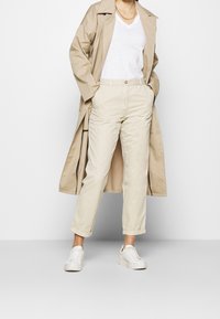 Cappotto trench beige sopra una t-shirt bianca, abbinato a chino beige chiaro e sneaker bianche. Presenta maniche arrotolate e una collana a catena.