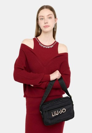 Borsa a tracolla in tessuto nero con logo dorato "LIU-JO", chiusura con zip e tracolla regolabile. Indossata con un maglione rosso caratterizzato da un design scollato sulle spalle.