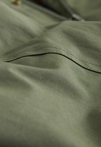 Olivgrüner Stoff mit glatter Textur und sichtbaren Nähdetails. Enthält subtile Falten, die auf ein geschichtetes Design hinweisen.