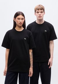 Μαύρα βαμβακερά μπλουζάκια με κοντά μανίκια, με ένα μικρό πράσινο λογότυπο στο στήθος, συνδυασμένα με σκούρο τζιν, που στέκονται μπροστά σε λευκό φόντο.