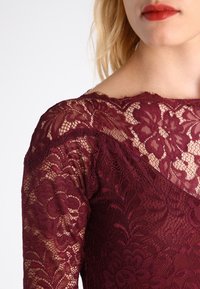 Burgunderfarbenes Spitzenkleid mit floralem Muster, das über einen Bootsausschnitt und durchsichtige lange Ärmel verfügt. Strukturierter, zarter Stoff mit filigranen Details.