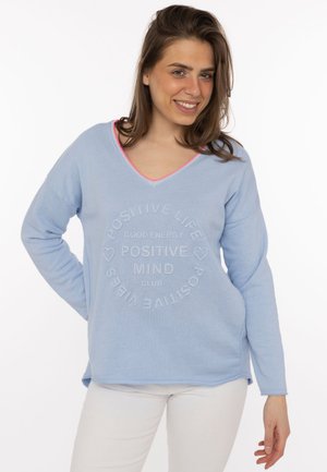 Lächelnde Frau mit braunen Haaren, die einen hellblauen Pullover mit dem Schriftzug „Positive Mind Club“ und weiße Hosen trägt, steht vor einem weißen Hintergrund.
