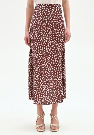 Donna con gonna midi marrone con macchie irregolari beige abbinata a sandali dorati con cinturini e tacco alto, in piedi davanti a uno sfondo bianco.