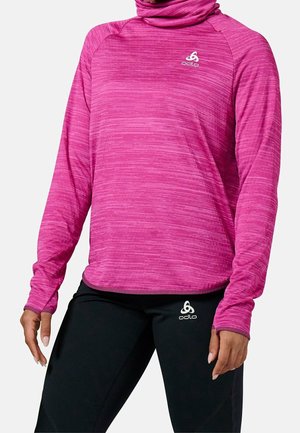 Vrouw draagt een roze lange mouwen sporttop met logo en zwarte leggings met logo, tegen een effen lichte achtergrond.
