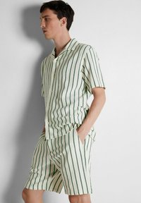 Šviesiai kreminės spalvos trumpų rankovių marškiniai su mygtukais ir šortai, padengti vertikaliomis žaliomis juostelėmis. Marškiniai turi laisvą siluetą ir tekstūruotą audinį.