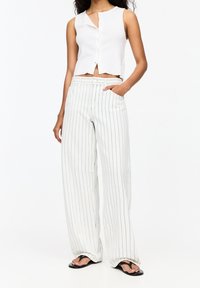 Haut blanc court sans manches avec fermeture à boutons, associé à un pantalon à jambes larges rayé taille haute en tissu léger. Tongs noires aux pieds.