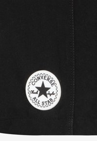 Svart stoff med en sirkulær hvit lapp med en stjerne og teksten "CONVERSE CHUCK TAYLOR ALL STAR". Glatt tekstur og sydd kant.