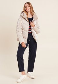 Doudoune beige avec une capuche en fourrure, associée à une chemise rayée, un pantalon navy et des baskets blanches sur un fond clair.