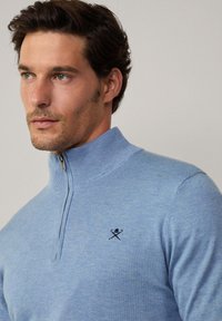 Svijetloplavi pleteni džemper s visokim ovratnikom i zatvaračem. Ima tamnoplavi logo na lijevoj strani prsnog koša. Mekana tekstura, prilagodljiv kroj.