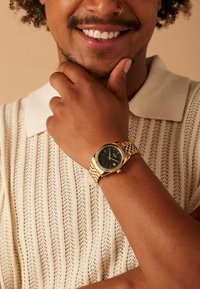 Gold wristwatch with a black face and textured bezel, worn on a light brown arm. The watch features a link strap and a patterned white knit shirt.
