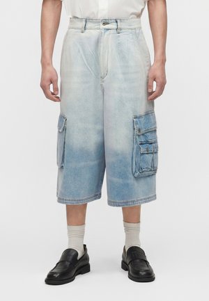 Άνδρας που φοράει ανοιχτό μπλε ξεθωριασμένο τζιν cargo σορτς μέχρι το γόνατο με μεγάλες τσέπες, λευκές κάλτσες και μαύρα μοκασίνια, στέκεται μπροστά σε λευκό φόντο.