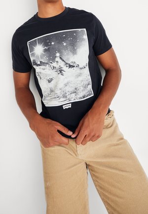 Person trägt ein schwarzes T-Shirt mit einer weißen Berg- und Sternenhimmel-Grafik und beigefarbenen Cordhosen, Hände ruhen nahe der Taille.