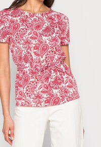 Mönstrad blus med röda och vita blommor, korta ärmar, rund hals och knytning i midjan; tillverkad av ett tunt, texturerat tyg.