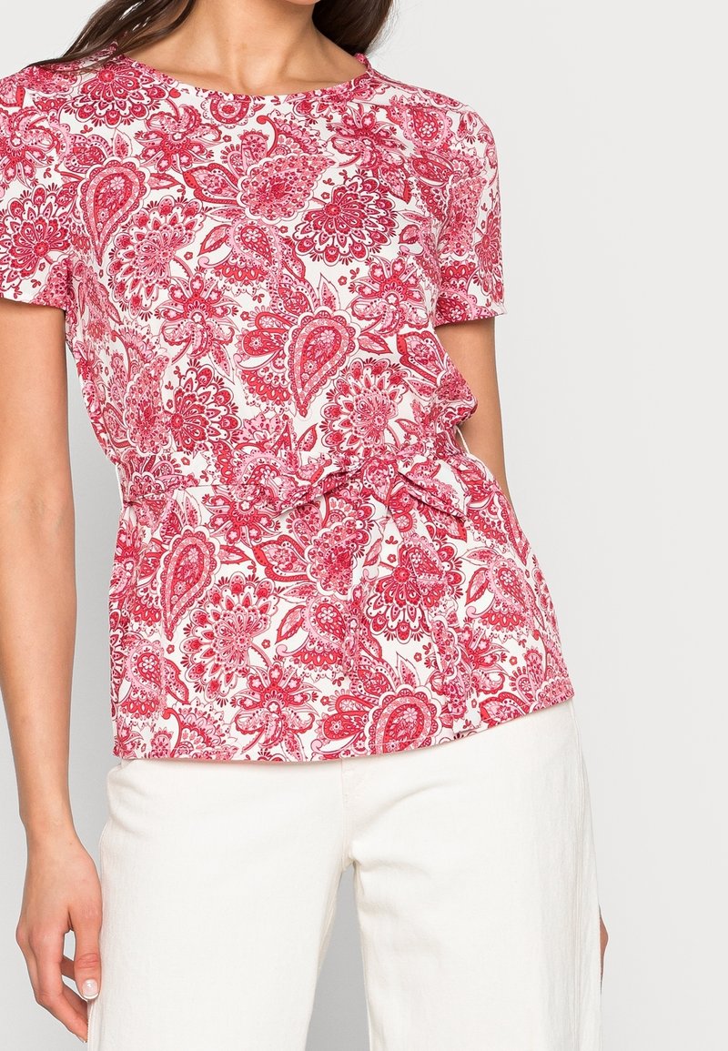 Mönstrad blus med röda och vita blommor, korta ärmar, rund hals och knytning i midjan; tillverkad av ett tunt, texturerat tyg.