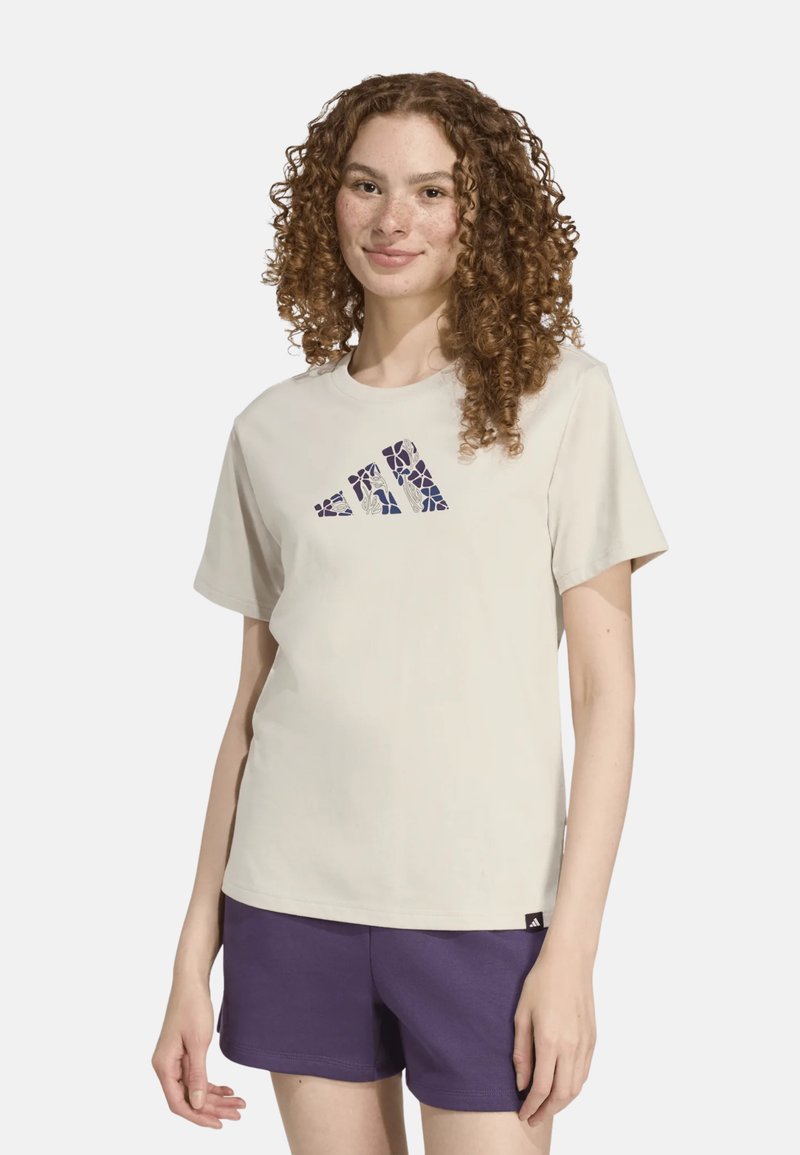 Camiseta de algodón beige con un logo gráfico morado. Mangas cortas, cuello redondo y etiqueta lateral. Combinada con pantalones cortos morados.