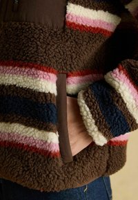 Fleece-Jacke mit braunen, pinken, roten und blauen Streifen; strukturierter, weicher Stoff; beinhaltet eine Tasche und einen Reißverschluss.