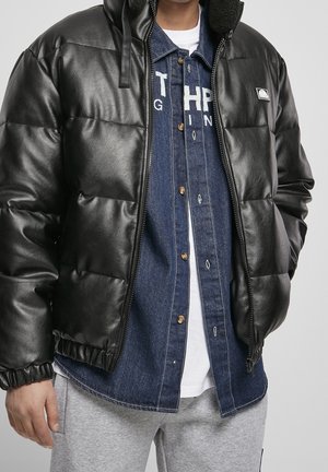 Mann in schwarzer Pufferjacke mit Kapuze, unknöpftem Jeanshemd über einem weißen T-Shirt und hellgrauen Sweatpants, steht mit entspannten Händen.