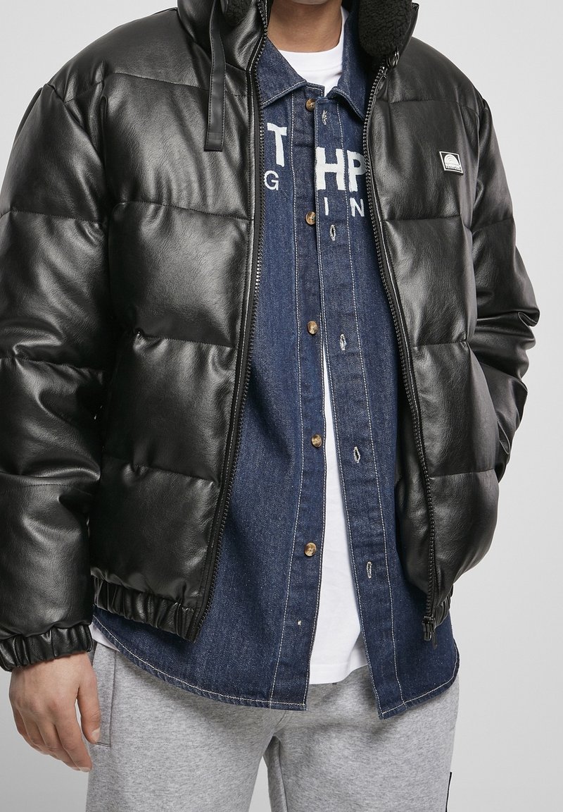 Man wearing black puffer jacket with hood, unbuttoned denim shirt over white tee, and light gray sweatpants, standing with hands relaxed.