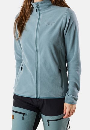 Frau trägt eine hellblaue Fleecejacke mit Reißverschluss und Seitentaschen sowie zweifarbige blaue und schwarze Outdoorhose mit Reißverschlusstaschen.