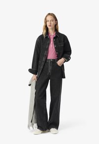 Giacca in denim nera e jeans neri a gamba larga abbinati, con una polo rosa. Sciarpa grigia appoggiata sul braccio. Vestibilità casual, hardware minimal.
