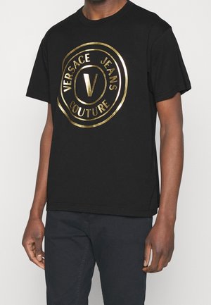 Osoba nosząca czarną koszulkę z krótkim rękawem, z dużym, złotym, okrągłym logo „Versace Jeans Couture” na piersi.