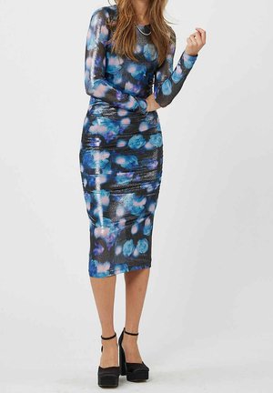 Mujer con un vestido entallado de manga larga hasta la rodilla con patrones abstractos en azul y rosa y tacones de plataforma negros.
