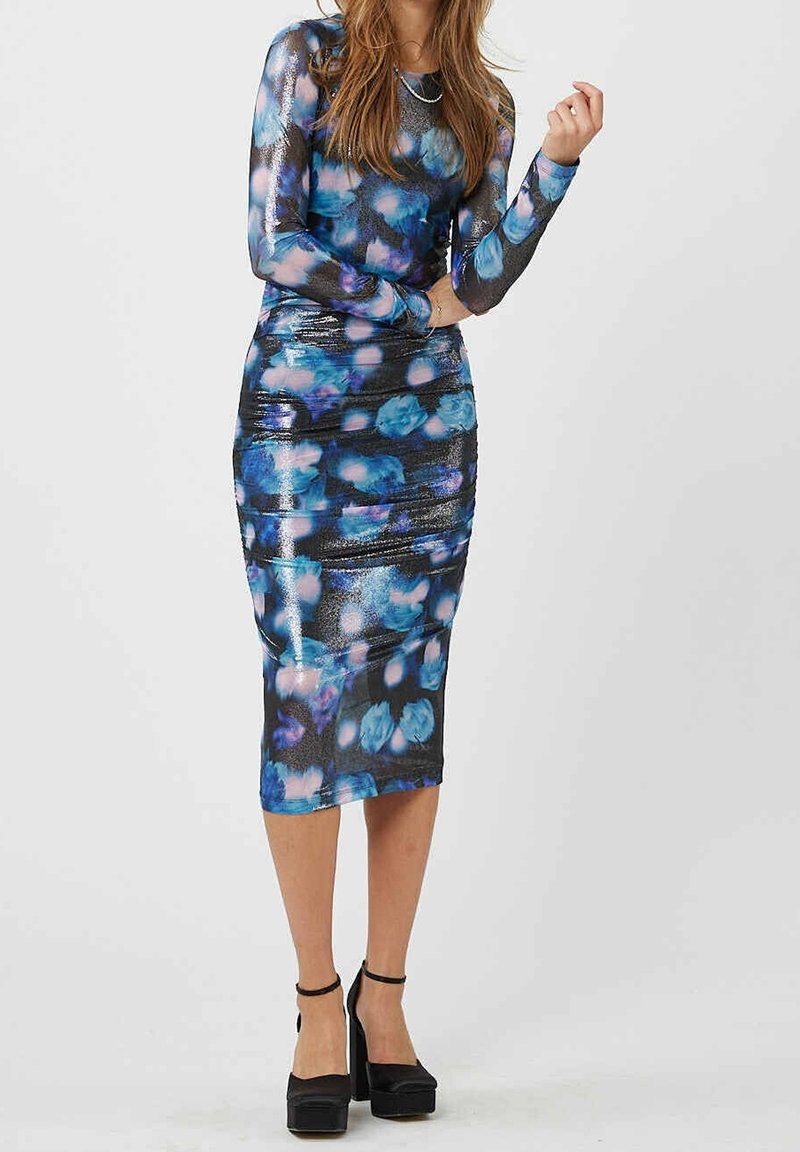 Femme portant une robe ajustée à manches longues, longueur au genou, avec des motifs abstraits bleus et roses et des talons compensés noirs.