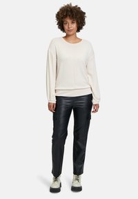 Helle beigefarbene Langarm-Weste mit Rundhalsausschnitt, kombiniert mit schwarzen Leder-Cargo-Hosen mit Seitentaschen und cremfarbenen Schnürstiefeln.