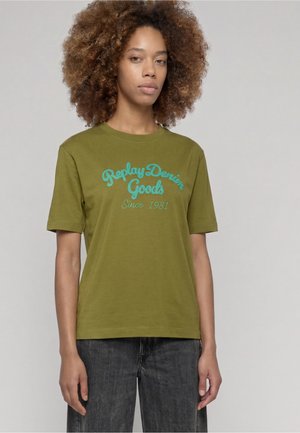 Femme aux cheveux bouclés portant un t-shirt vert olive avec un texte bleu "Replay Denim Goods Since 1981" et un jean noir sur un fond uni.