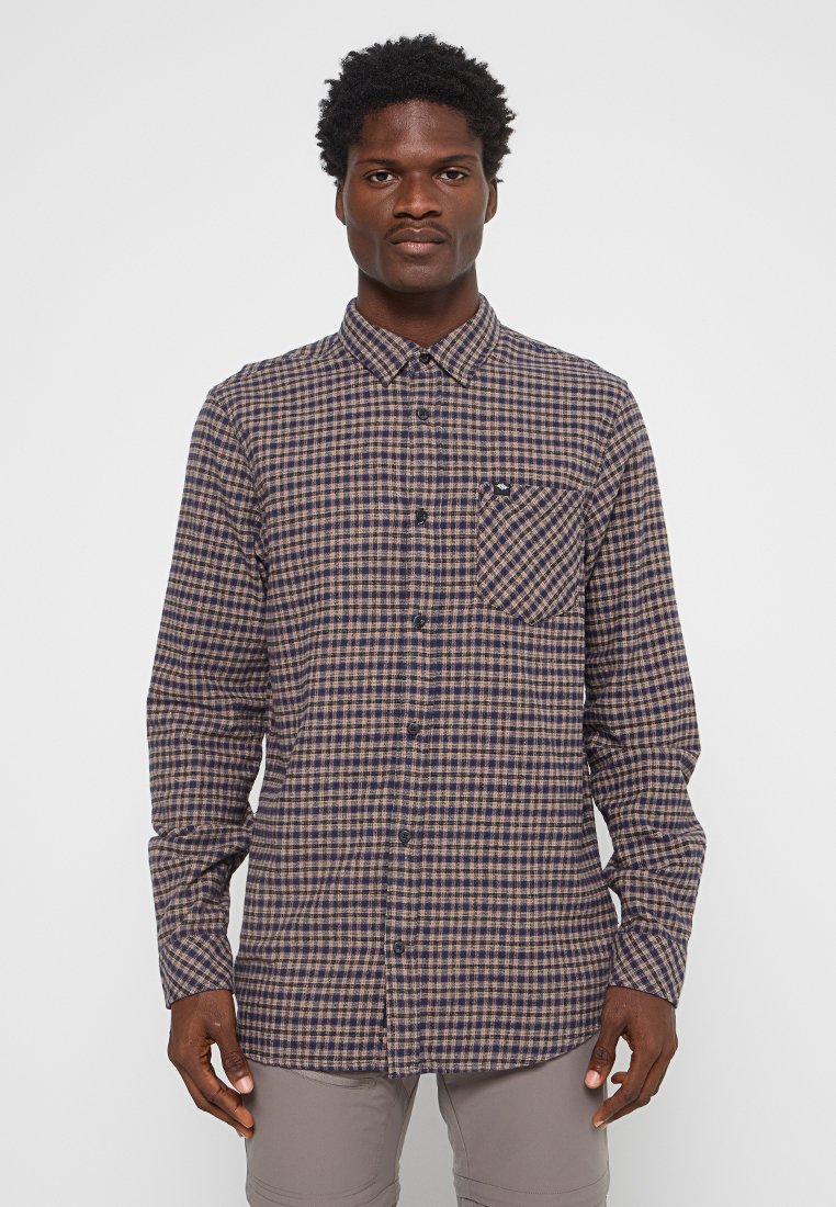 Homme portant une chemise à carreaux marron et bleu marine à manches longues avec poche avant, se tenant devant un fond clair uni.