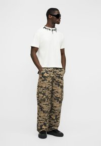 Άνδρας φοράει μαύρα γυαλιά ηλίου, λευκό μπλουζάκι με μαύρο κείμενο στον γιακά, παραλλαγής παντελόνια cargo και μαύρα παπούτσια, στέκεται με τα χέρια στις τσέπες.