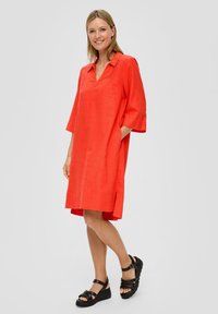Rotes Leinenkleid mit V-Ausschnitt, Dreiviertelärmeln und seitlichen Taschen. Der Saum ist hinten etwas länger. Kombiniert mit schwarzen Plateausandalen.