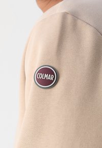Beige sweatshirt met een gladde textuur, voorzien van een rond Colmar logo-embleem in bordeauxrood en zilver op de linkerschouder.