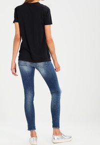 Žena oblečená v černém tričku s krátkým rukávem, vybledlých modrých skinny jeans a stříbrných slip-on botách, stojící zády k divákovi.