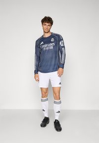 Maglia da calcio a maniche lunghe di colore blu scuro con accenti bianchi e logo, abbinata a pantaloni bianchi e scarpini neri. Calze a righe completano il look.