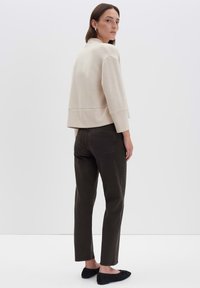 Beige verkürzter Pullover mit weiten Ärmeln, kombiniert mit dunkelgrünen, geraden Hosen und schwarzen flachen Schuhen, vor einem einfarbigen Hintergrund.