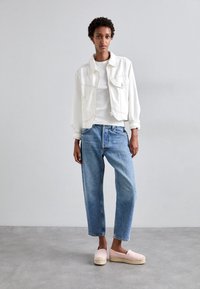 Chaqueta de mezclilla blanca con dos bolsillos frontales, diseño ajustado, combinada con jeans sueltos de color azul claro y alpargatas rosas. Fondo plano.