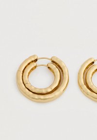 Pár kovových zlatých náušníc v tvare kruhu s textúrou a sústredným, kladivkovým dizajnom na bielom pozadí.