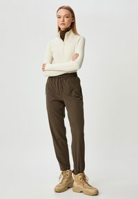 Cremfarbener Reißverschlussoberteil, braune Hose mit elastischem Bund und sandfarbene Wanderstiefel. Glatter Stoff, lockere Passform, funktionale Taschen und strukturierte Details an den Stiefeln.