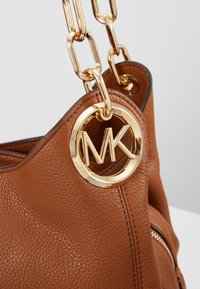 Sac à main en cuir marron texturé avec bandoulière chaîne dorée et breloque ronde dorée avec logo MK attachée près du bord supérieur.