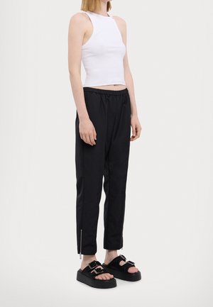 Kobieta ubrana w białą, bezrękawnikową bluzkę typu crop top, czarne, zwężane spodnie z suwakami przy kostkach oraz czarne platformowe sandały z klamrami.